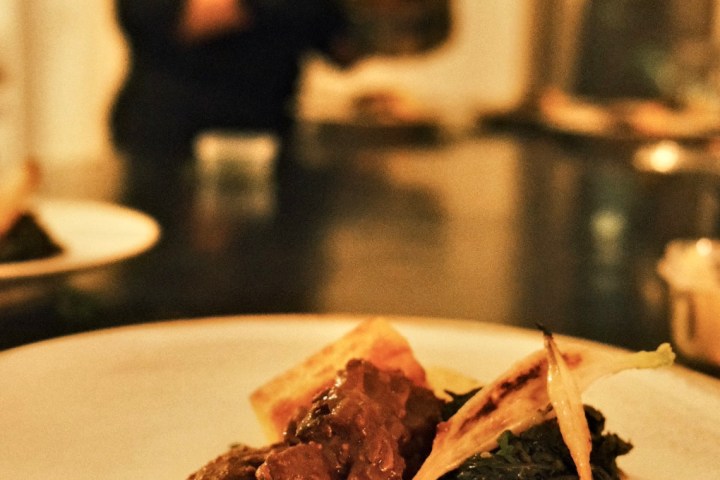 Gourmet dish with meat, greens, and garnish on white plate; blurred chef in background.
