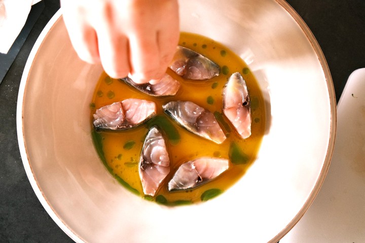 Hand seasoning raw fish slices in a bowl with yellow and green liquid.