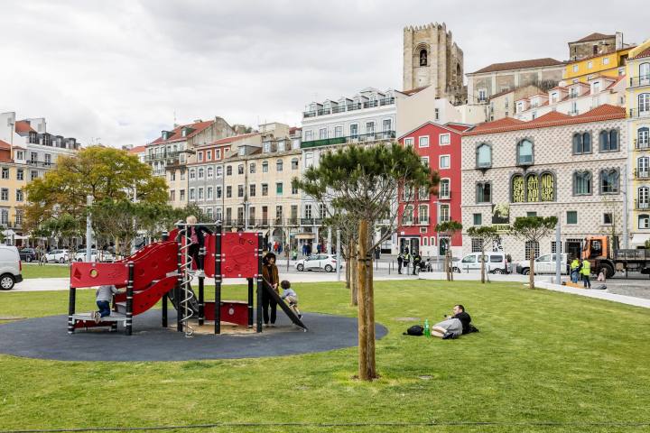 Children playing on a red playground in a city park with historic buildings in the background.