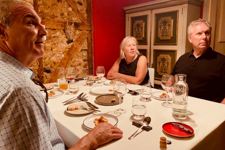 Three people seated at a dining table with plates and wine glasses, in a rustic room with exposed brick.