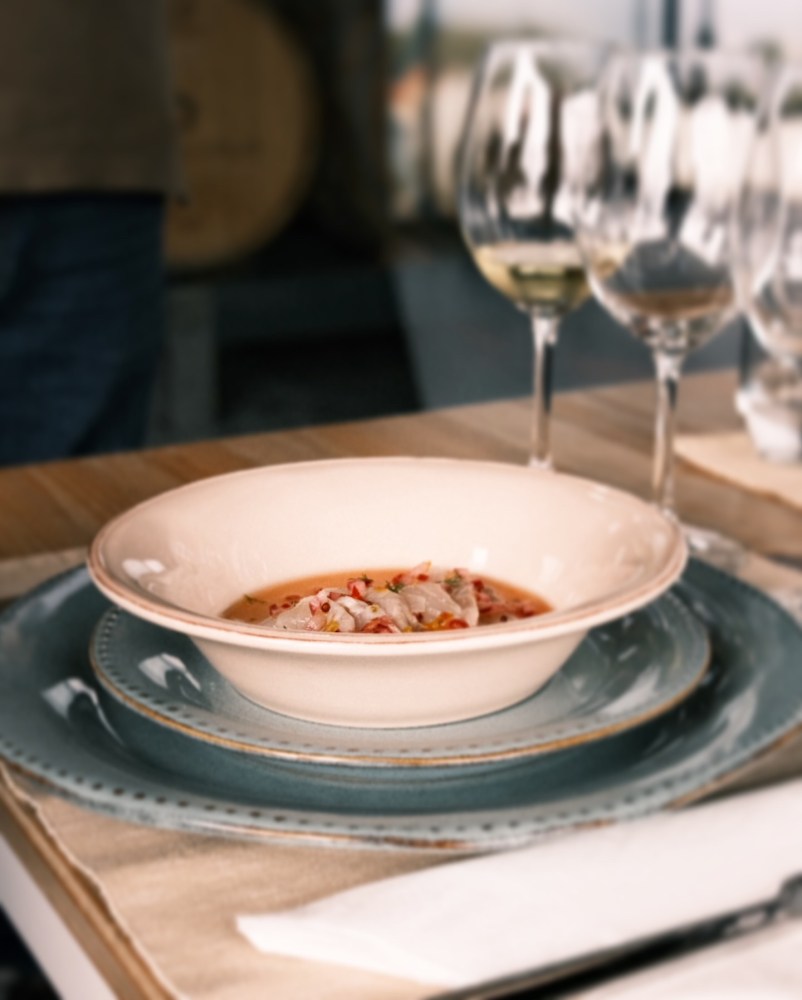 Dish of seafood soup on stacked plates with wine glasses in the background.