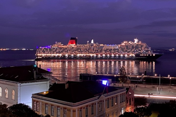 Lit cruise ship on water at night with city buildings in foreground.