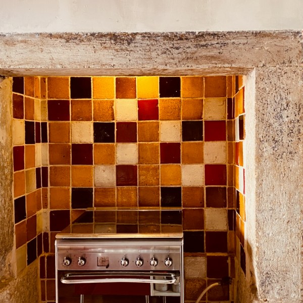 Colorful tiled kitchen backsplash with a stainless steel stove and wooden shelves with kitchen utensils.