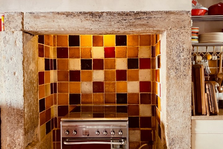 Colorful tiled kitchen backsplash with a stainless steel stove and wooden shelves with kitchen utensils.