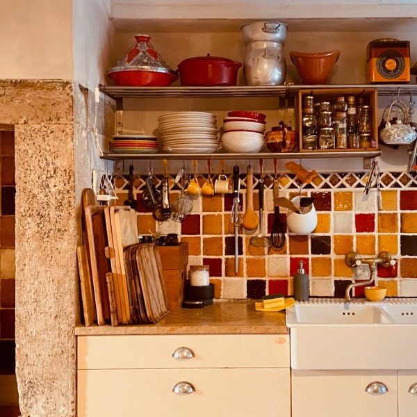 Rustic kitchen with tiled backsplash, shelves with dishes and spices, hanging utensils, and cutting boards.