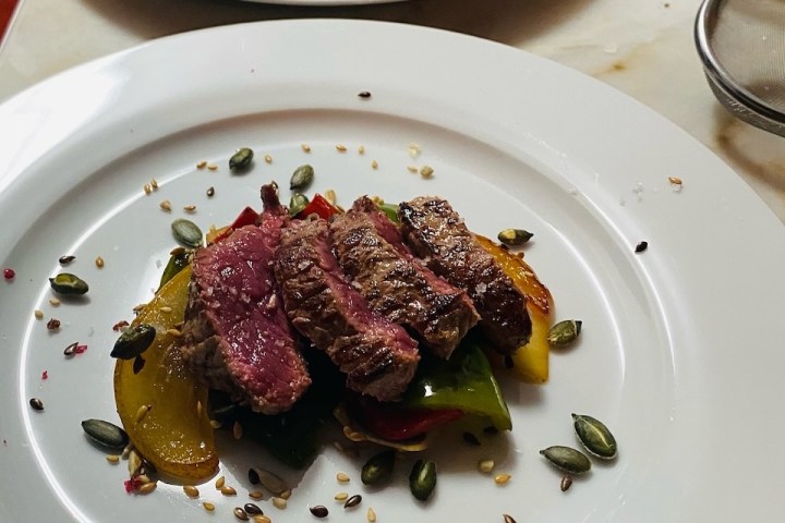 Two plates with sliced steak on roasted vegetables, garnished with seeds on a white table.