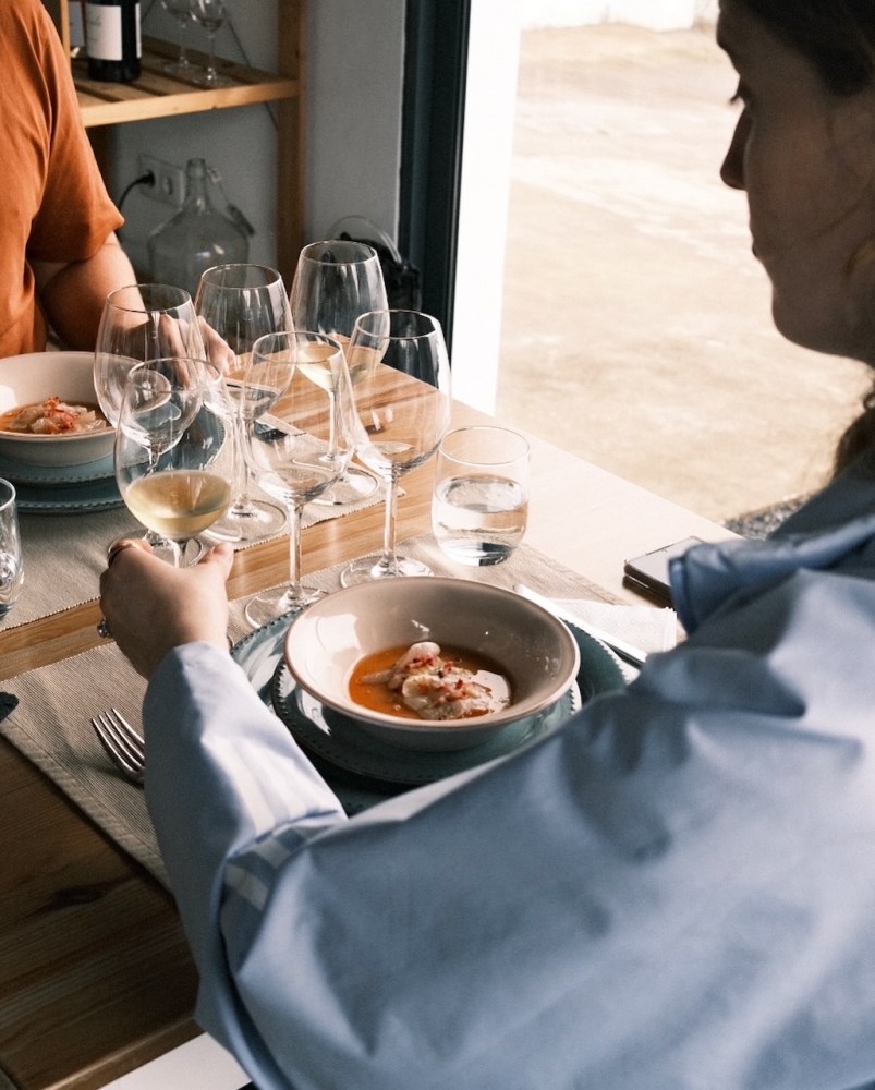 People dining with bowls of food and glasses of white wine at a wooden table.