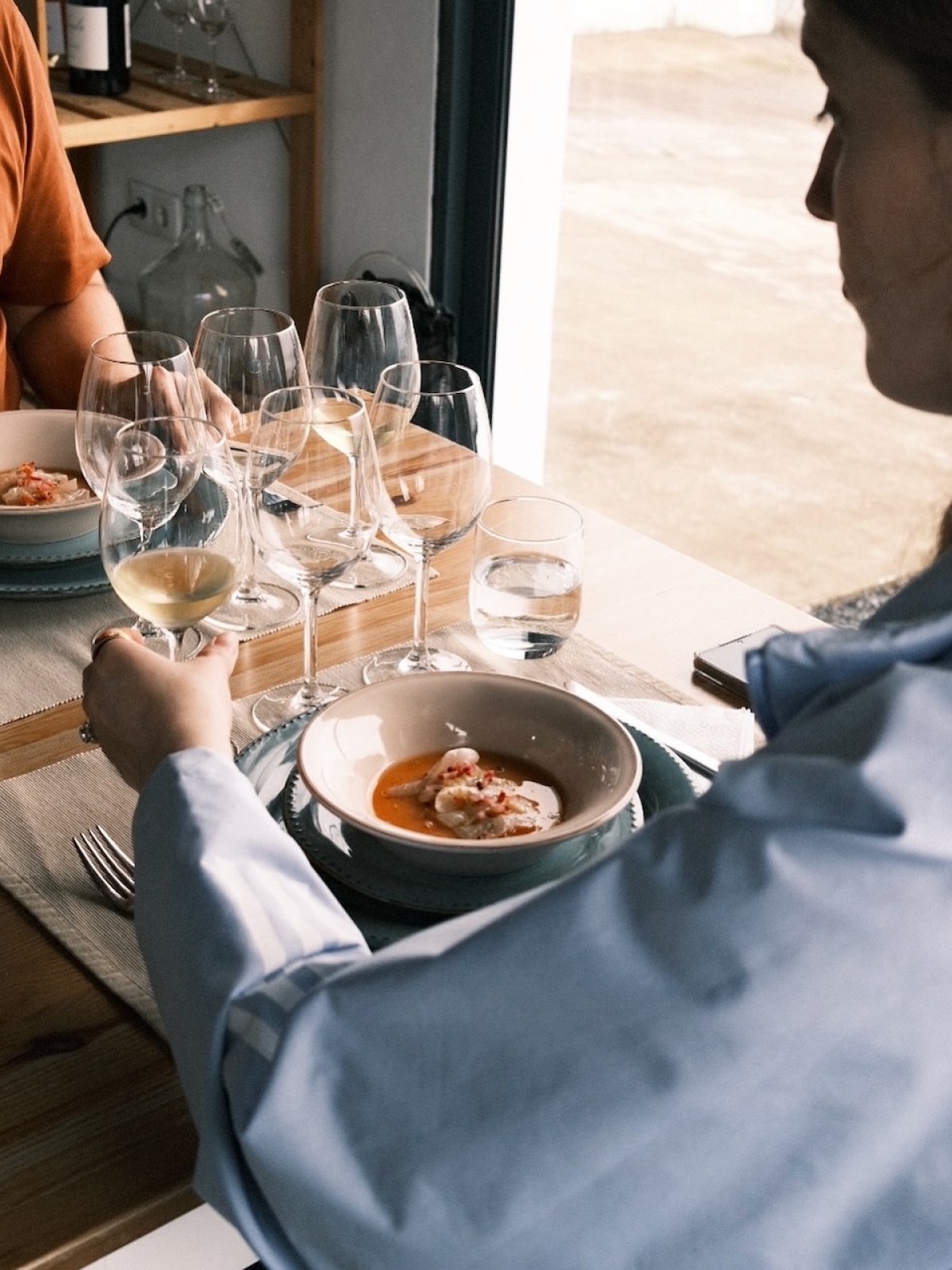 People dining with bowls of food and glasses of white wine at a wooden table.