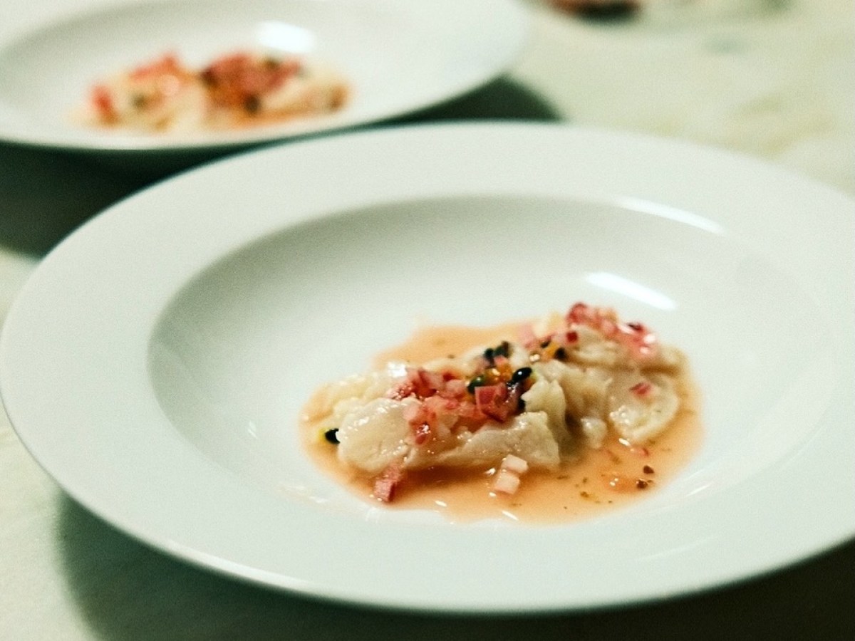 White plate with gourmet fish dish topped with sauce and garnishes on a table.