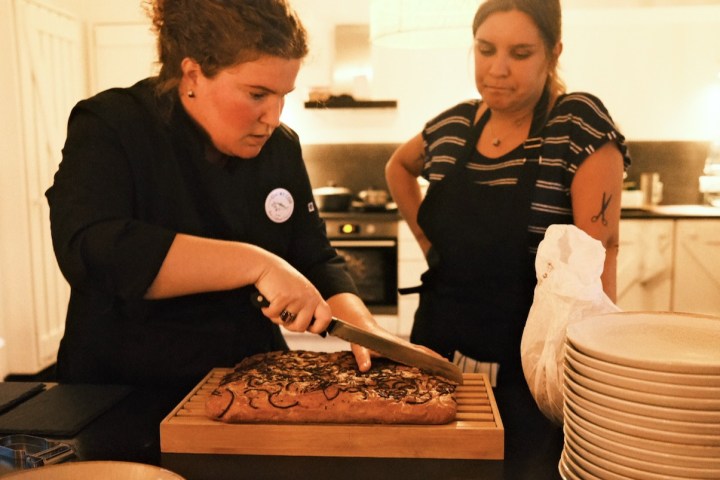 Two women in a kitchen, one slicing bread, with stacked plates nearby.