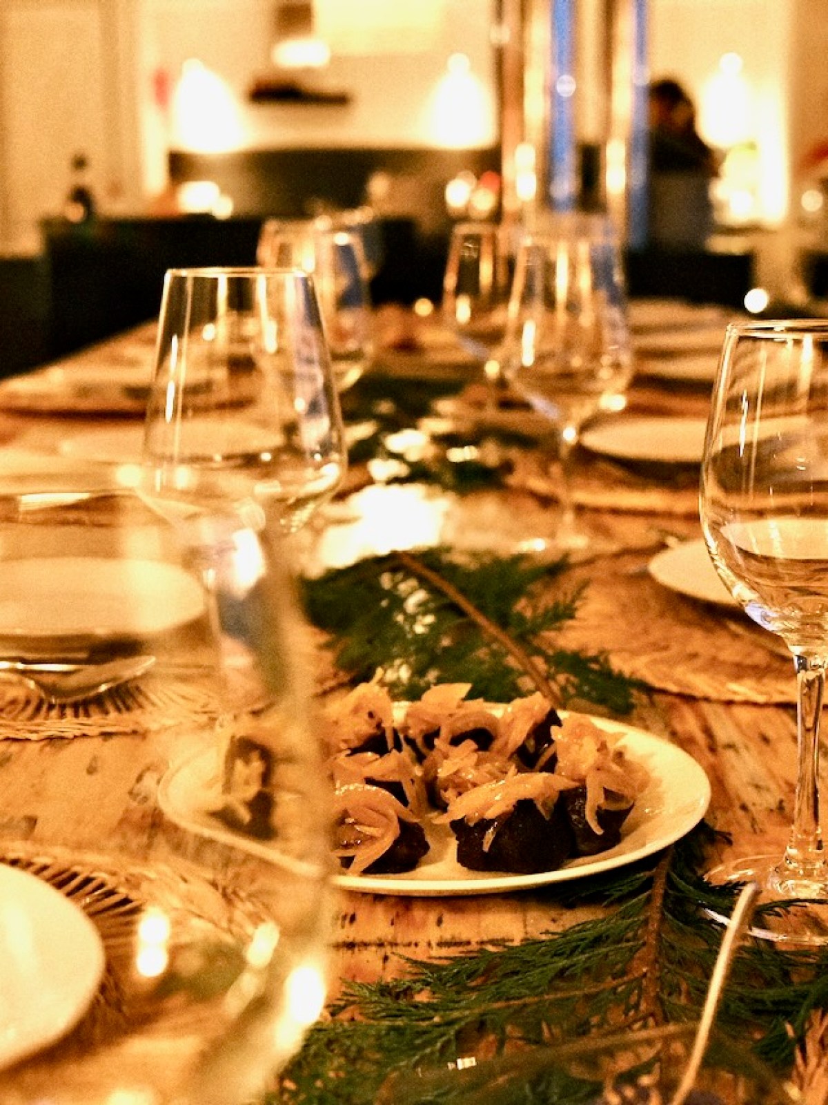 A set dining table with wine glasses, plates, and a centerpiece of greenery and small appetizers.