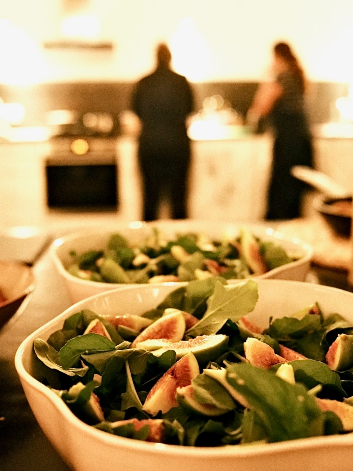 Bowls of fresh salad with figs in foreground, blurred kitchen with two people in background.