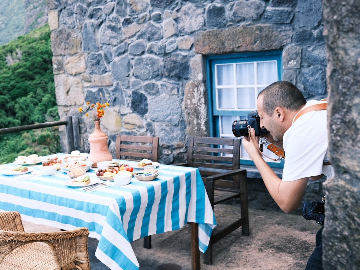 Photographer taking pictures of food on a table with striped cloth outdoors by a stone building.