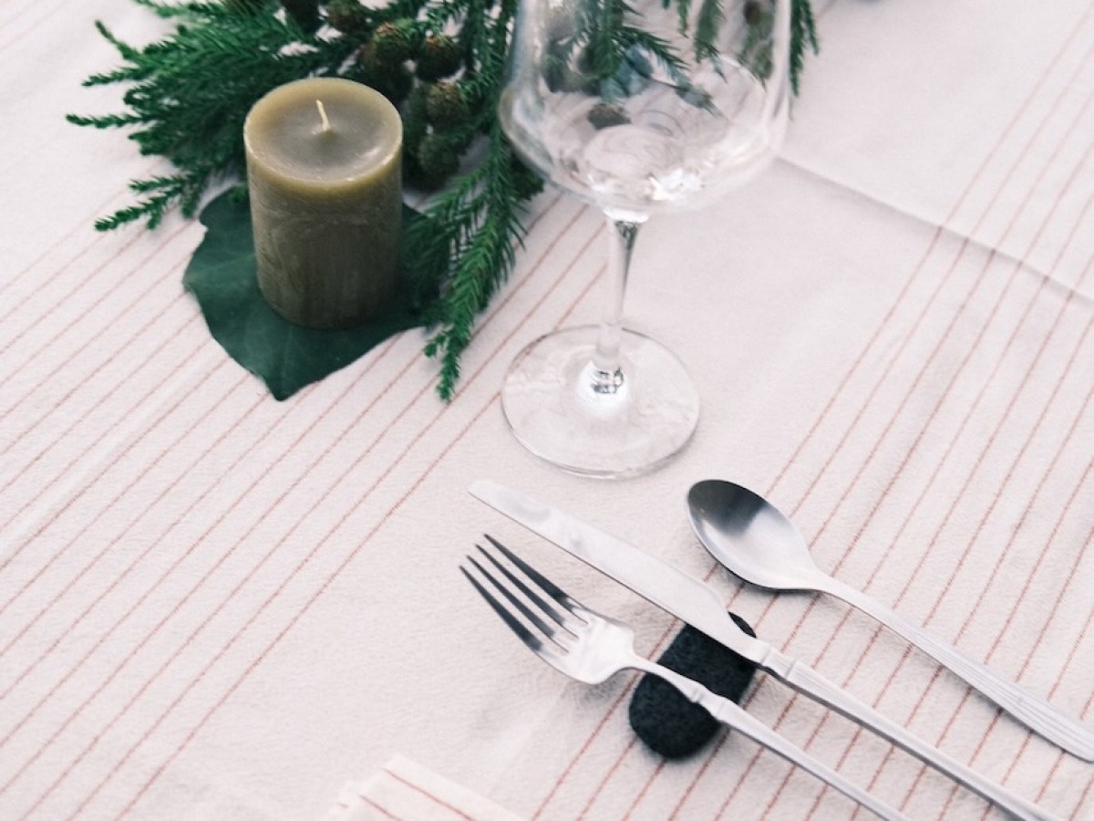 Table setting with greenery, candles, wine glass, and cutlery on white cloth.