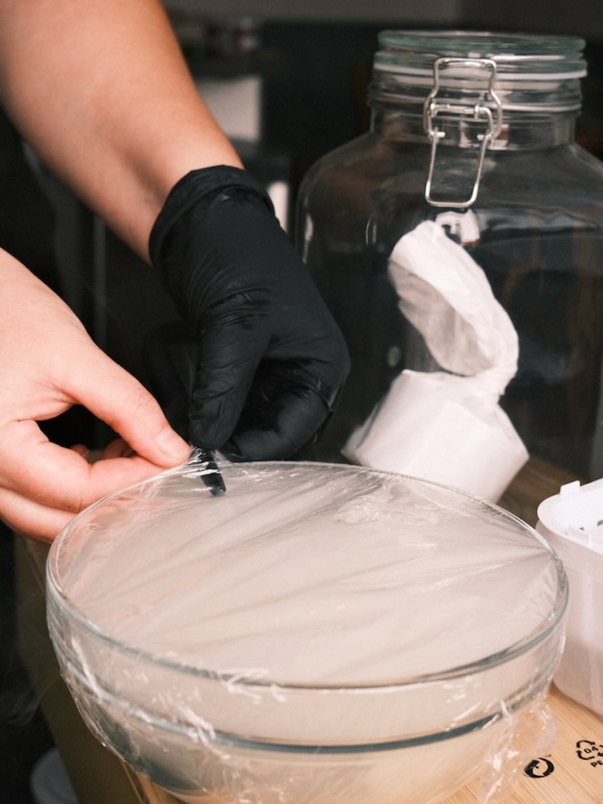 Person wrapping a glass bowl with plastic wrap, kitchen jars visible nearby.
