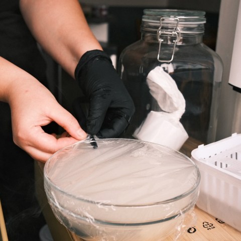 Person wrapping a glass bowl with plastic wrap, kitchen jars visible nearby.