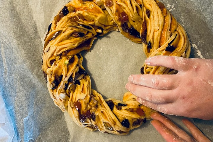 Hands shaping a braided fruit bread wreath on parchment paper.