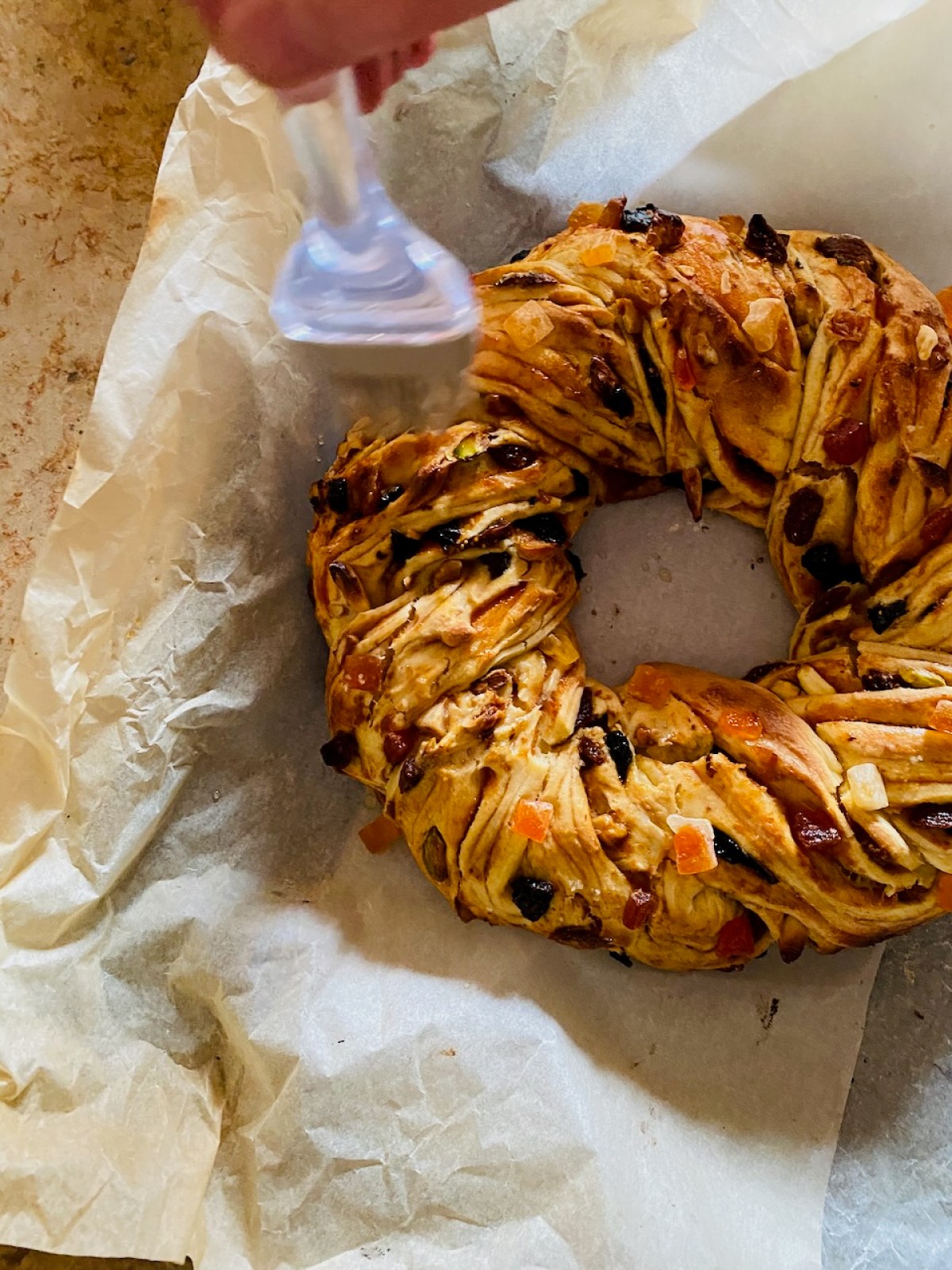 Person brushes glaze on braided fruit bread wreath on parchment paper.