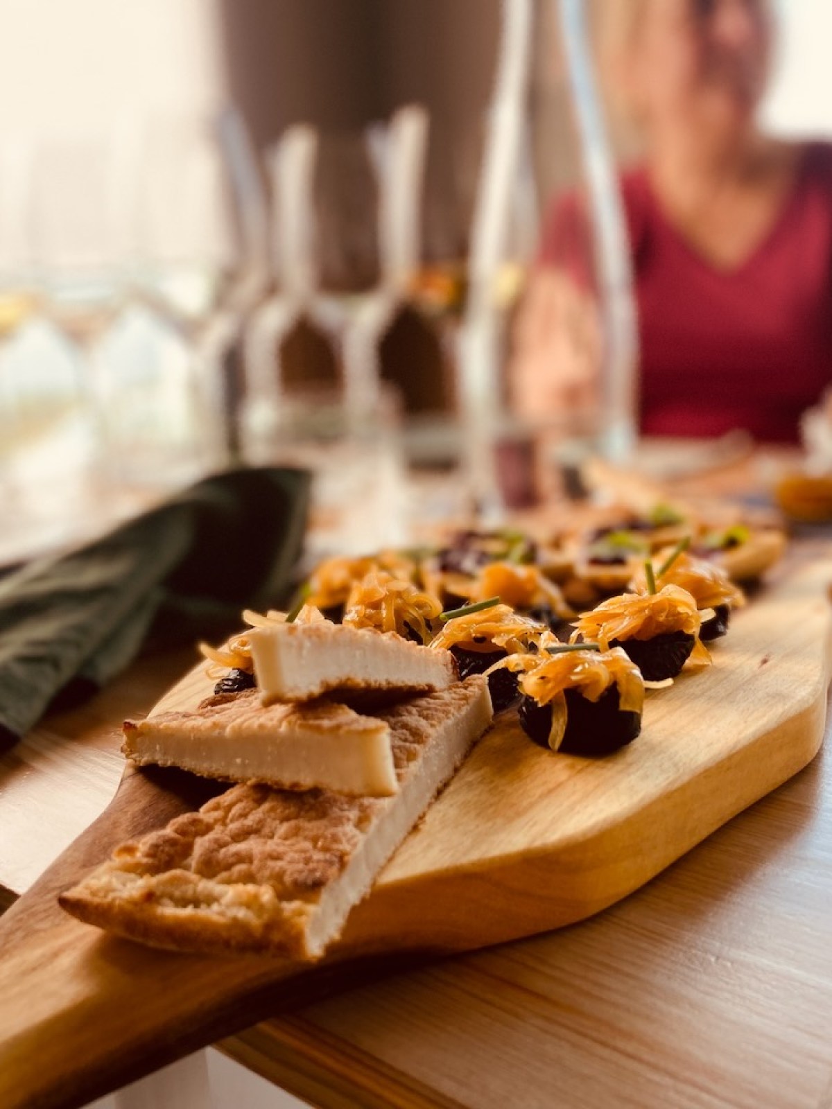 Cheese and appetizers on a wooden board with blurred diners in the background.