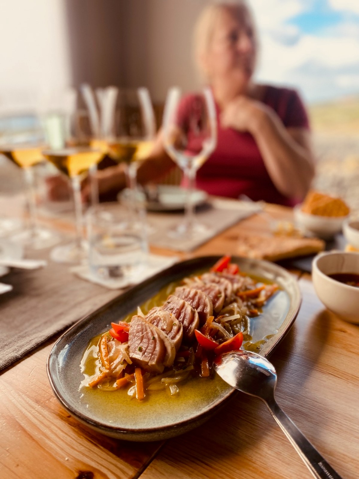 Plate of meat and vegetables on wooden table, with person and wine glasses in the background.