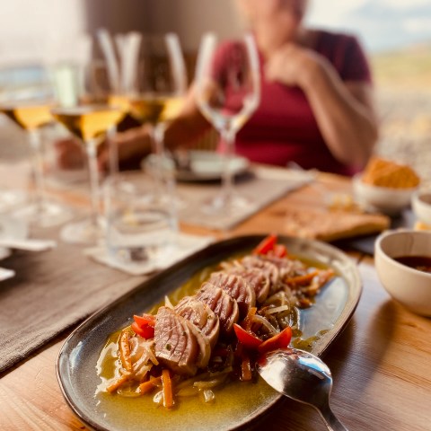 Plate of meat and vegetables on wooden table, with person and wine glasses in the background.