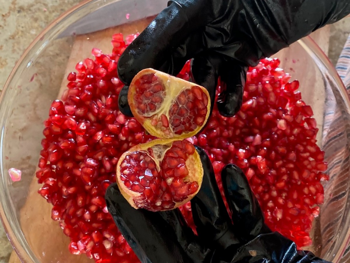 Hands in black gloves holding pomegranate halves over a bowl of seeds.