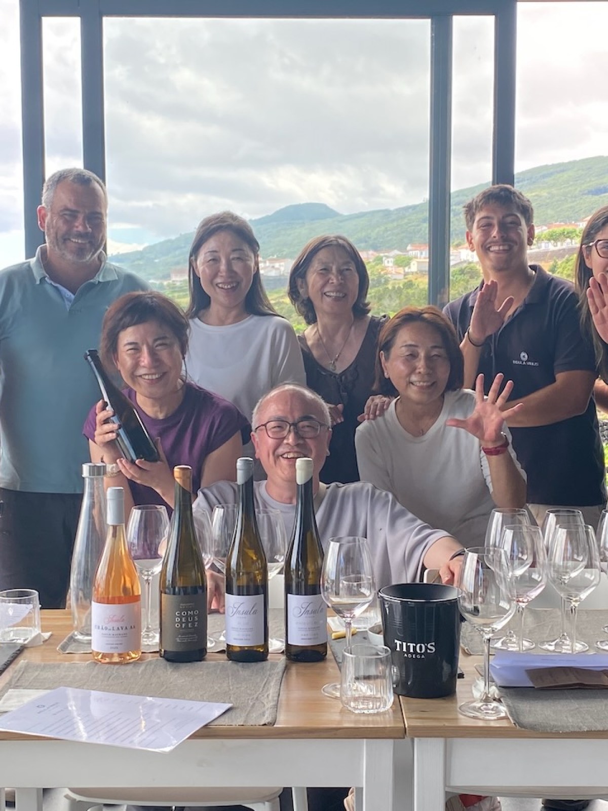 A group of people smiling at a wine tasting with wine bottles and glasses on the table.
