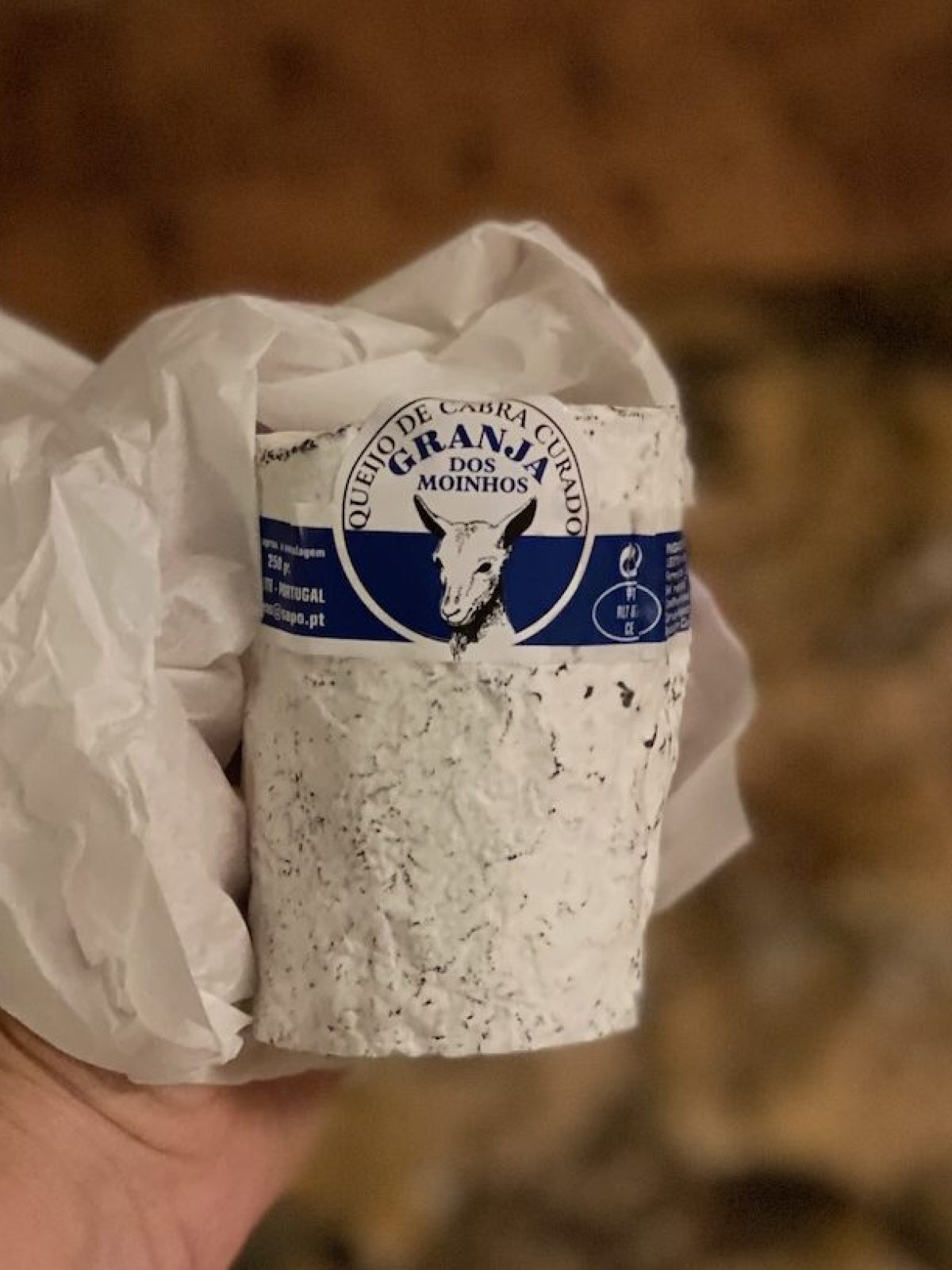 Hand holding a round, wrapped cheese labeled 'GRANJA DOS MOINHOS' against a blurred background.