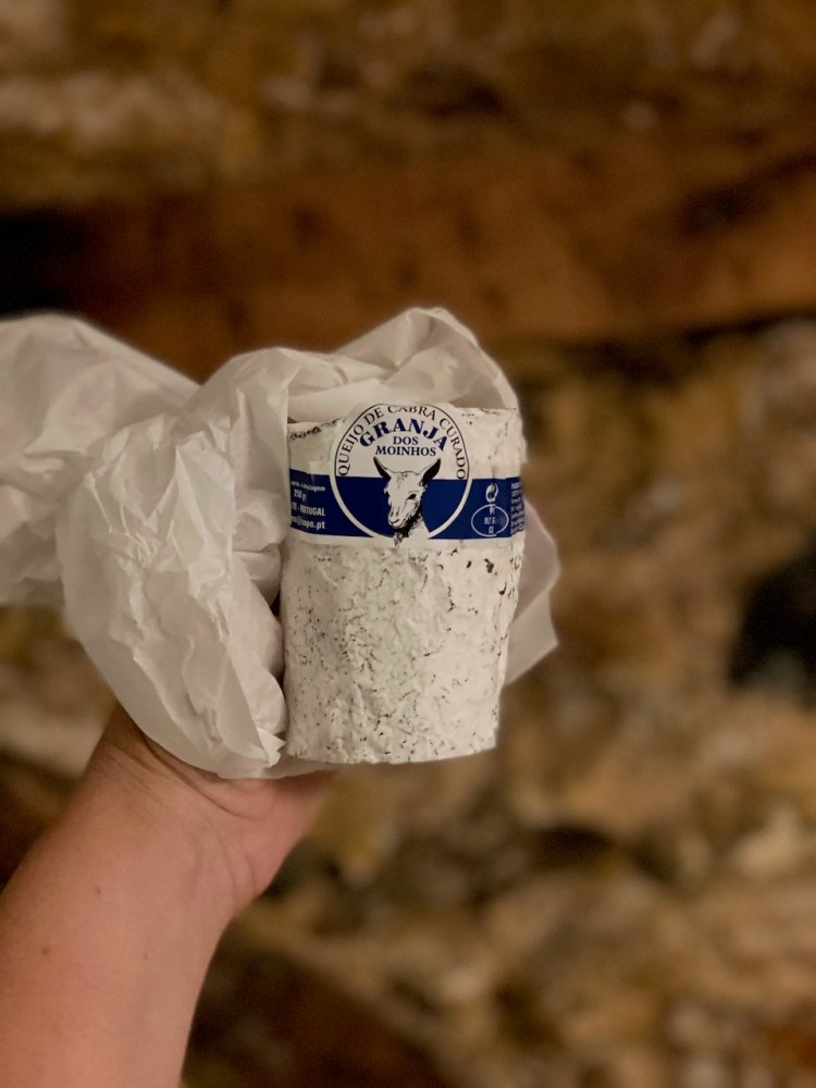 Hand holding a round, wrapped cheese labeled 'GRANJA DOS MOINHOS' against a blurred background.