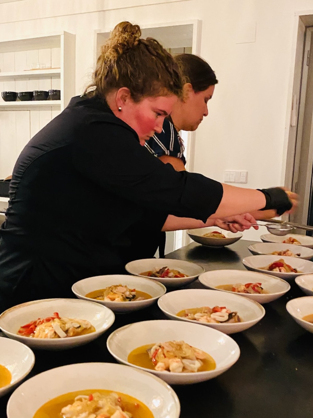 Two chefs plating soup dishes in a kitchen with several bowls on a counter.