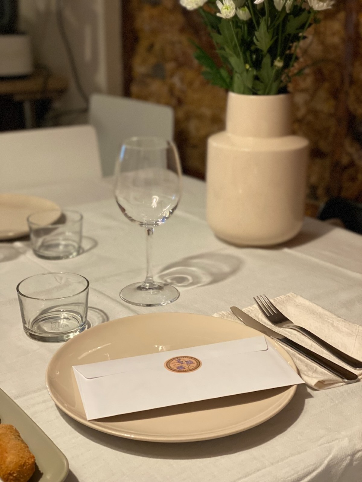 Table setting with envelope on plate, glass, cutlery, and vase with flowers.