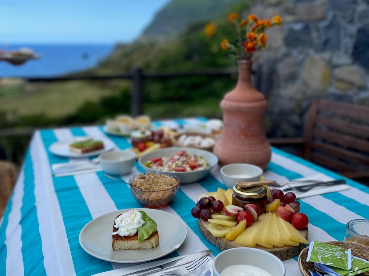 Outdoor table with breakfast food, fruit platter, and vase, overlooking ocean and mountains.