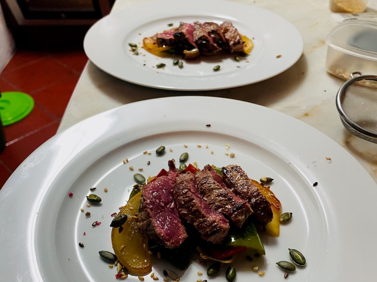 Sliced steak on roasted vegetables, garnished with seeds on two white plates in a kitchen setting.