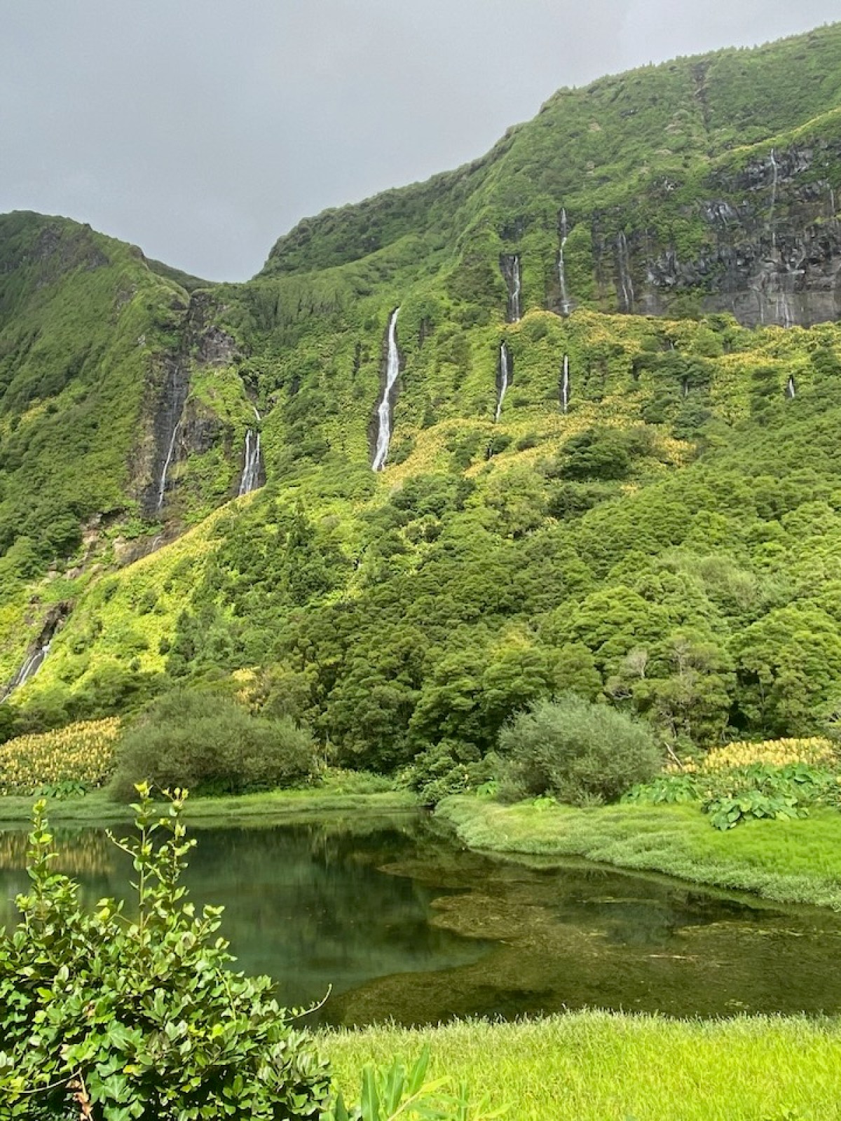 Lush green mountainside with multiple waterfalls and a reflecting pond below.