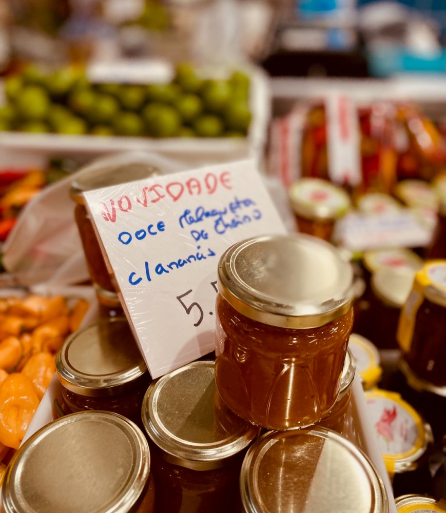Jars of jam with a handwritten sign on a market stall, assorted produce in background.