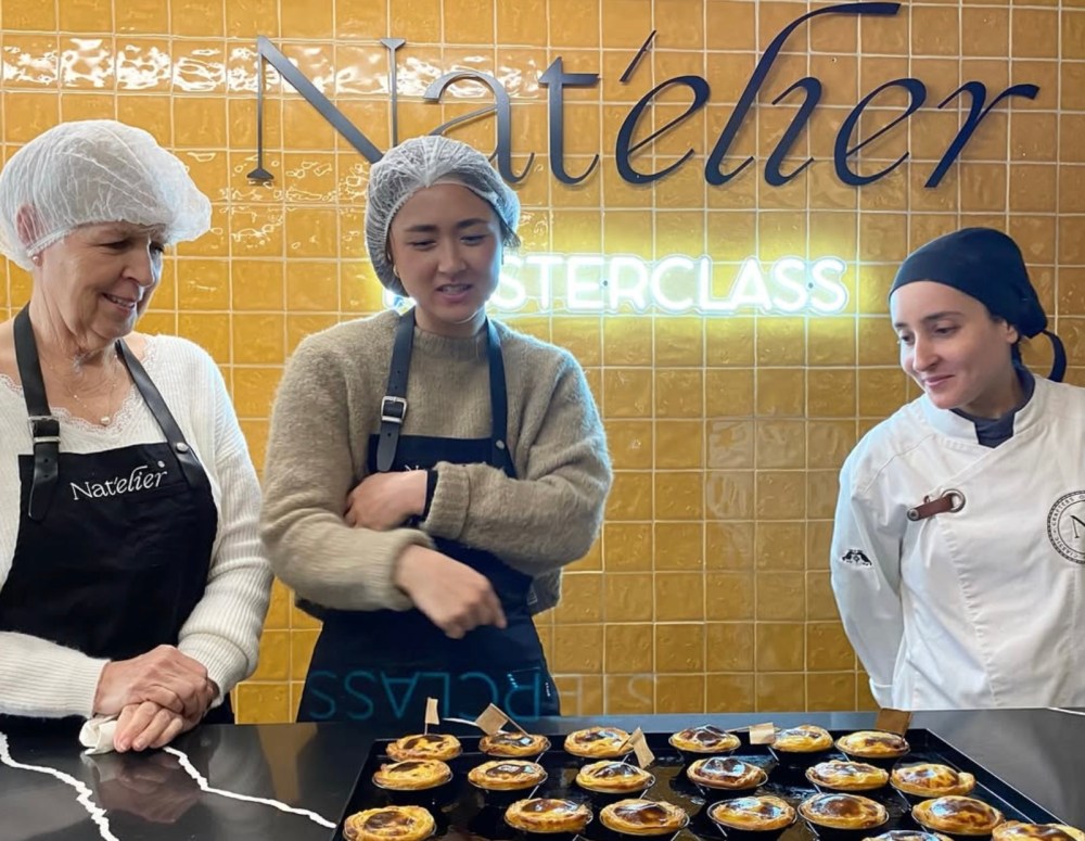 Three people in aprons and hairnets stand behind trays of pastries in a kitchen.