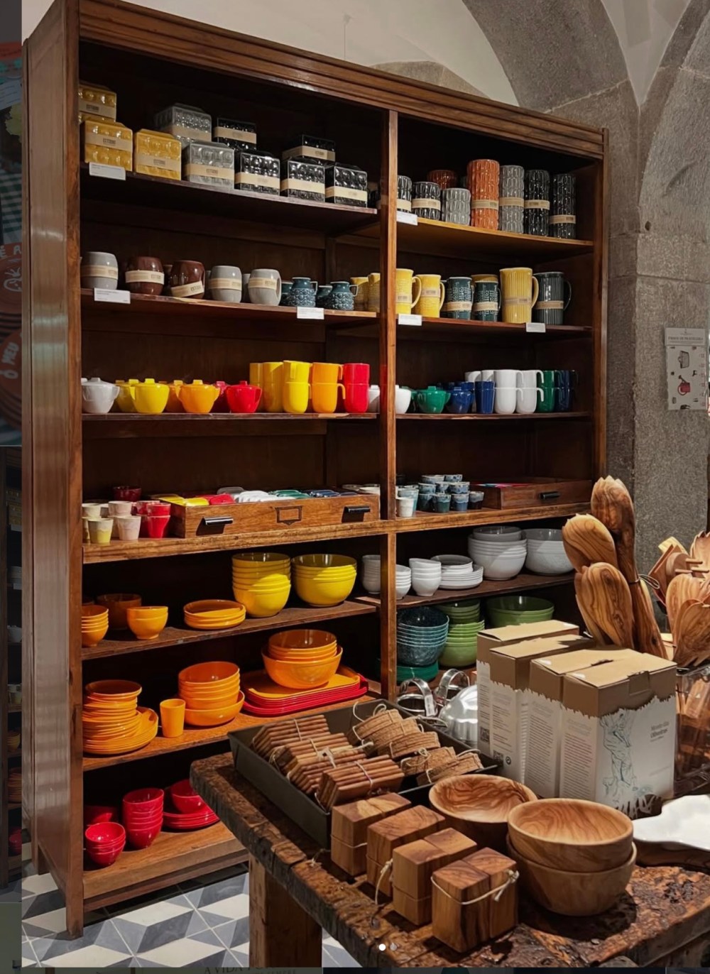 Wooden shelves display colorful mugs and bowls, with wooden utensils and boxes on a table in the foreground.
