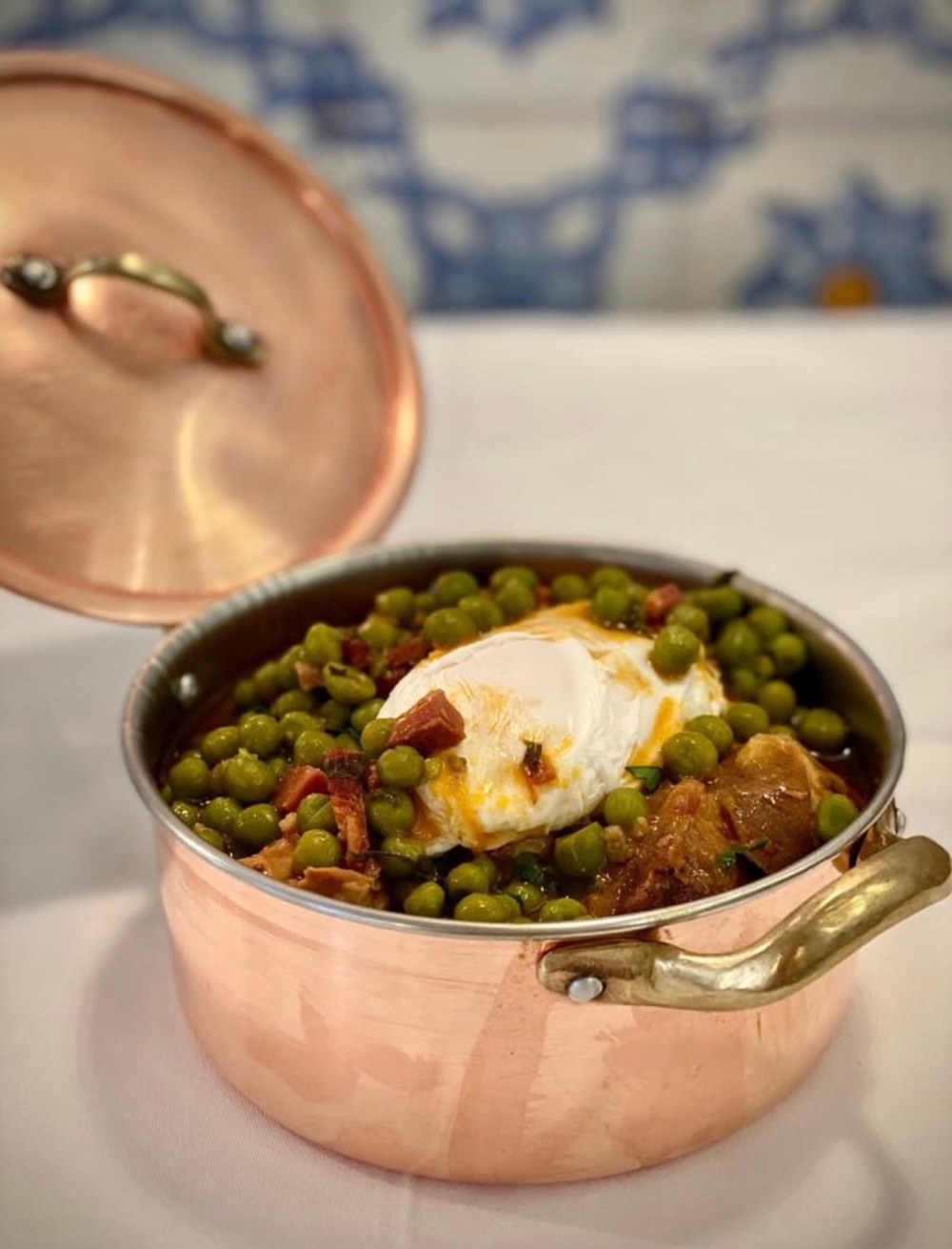 Copper pot with peas, meat, and a poached egg, open lid beside it on a white cloth background.