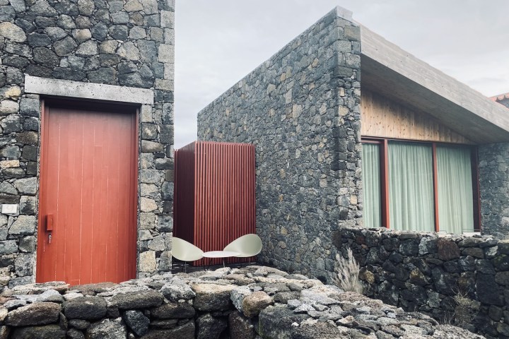 Stone building with red door and window, features modern design and outdoor chair with cloudy sky.