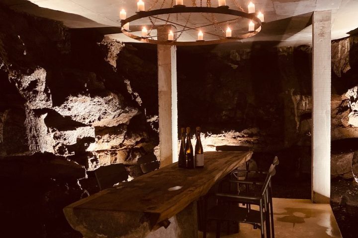 Rustic dining table with wine bottles in a dimly lit cave, chandelier above.