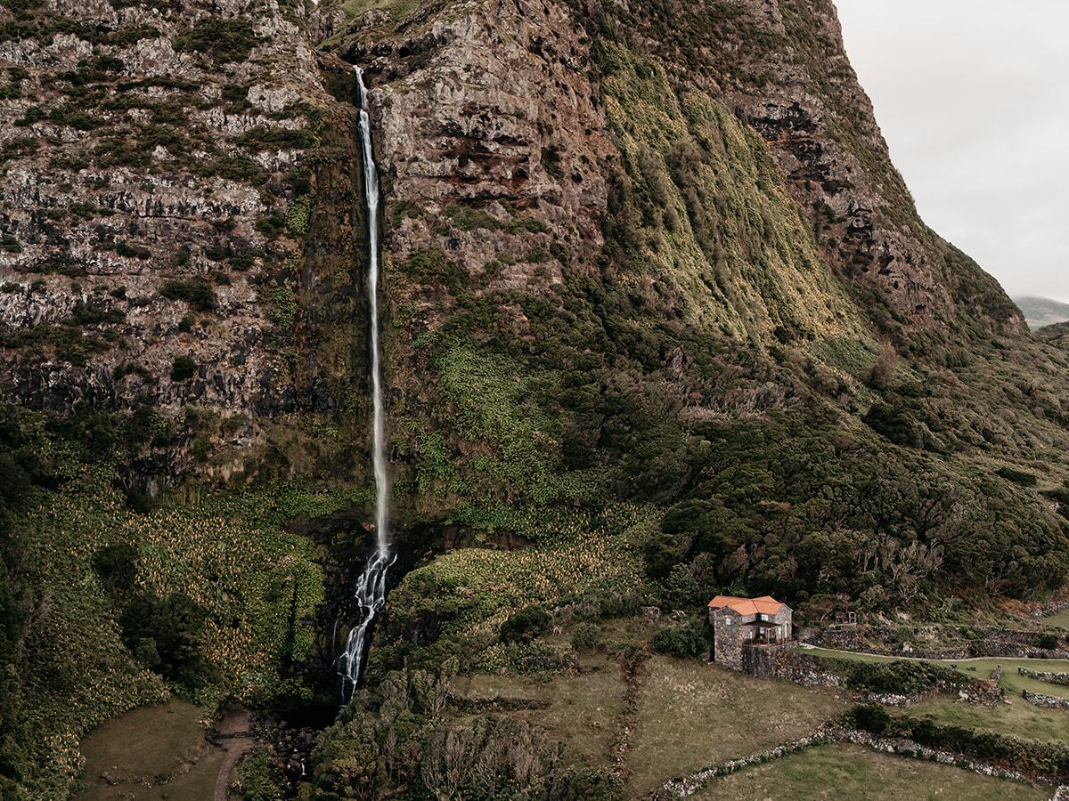 Tall waterfall cascading down a rocky cliff with greenery, near a small stone building with a red roof.