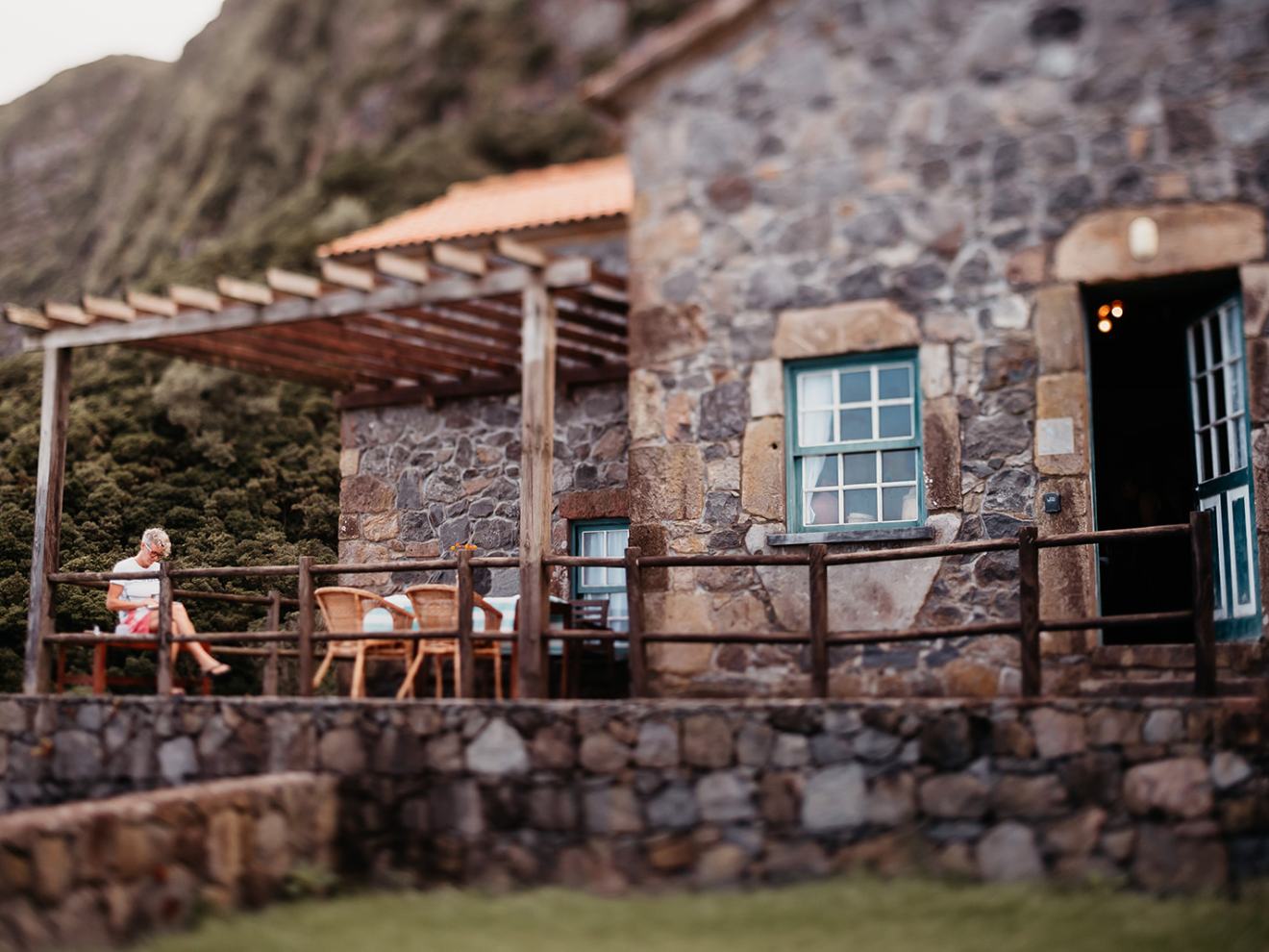 Person sitting on stone patio of rustic house with wooden pergola, surrounded by greenery.