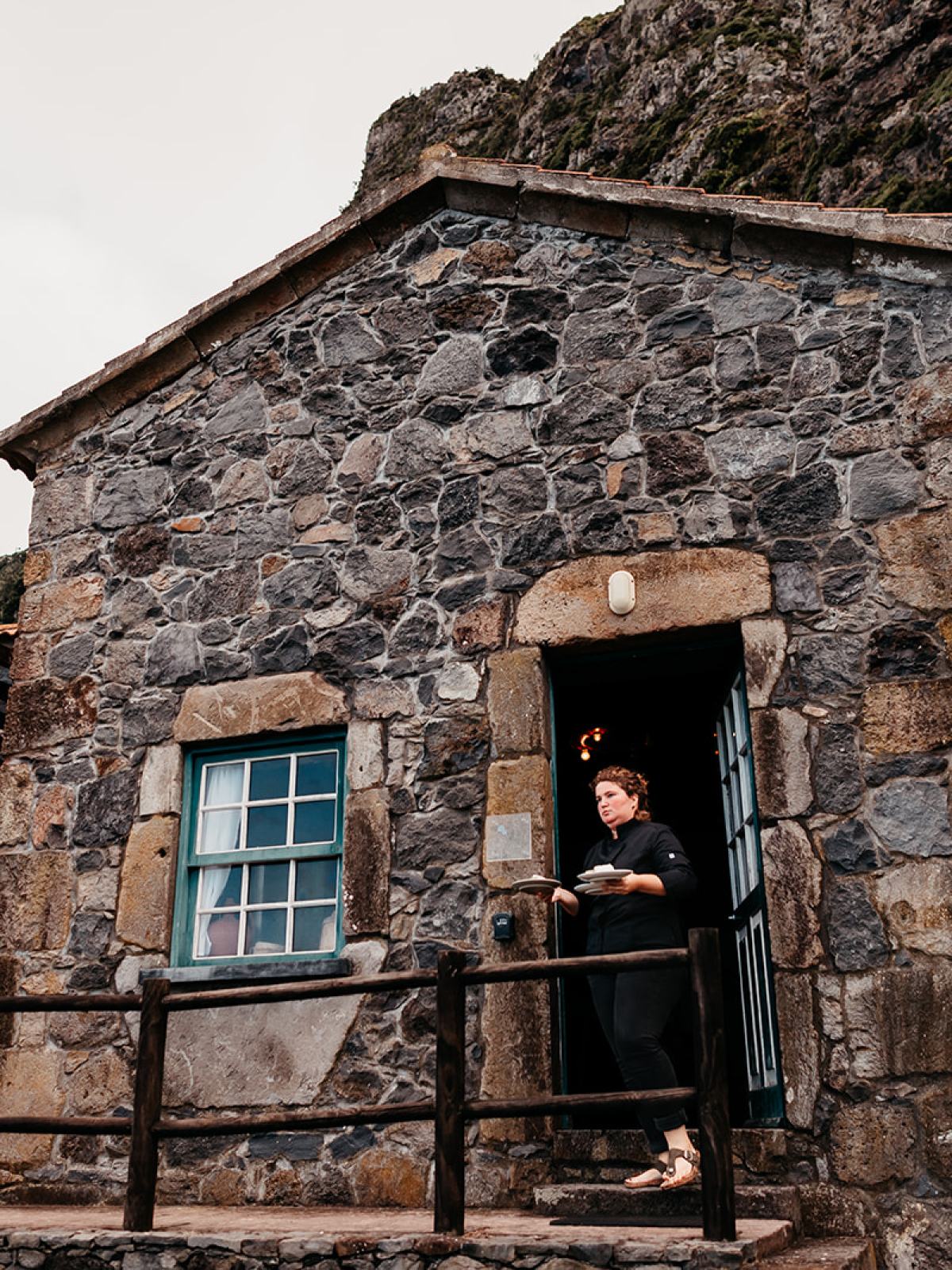 Stone building with a person standing in the doorway, near a table set outside under a pergola.