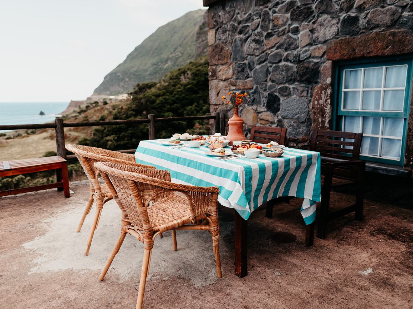Outdoor table with striped cloth, food, and chairs by a stone house overlooking the sea and hills.