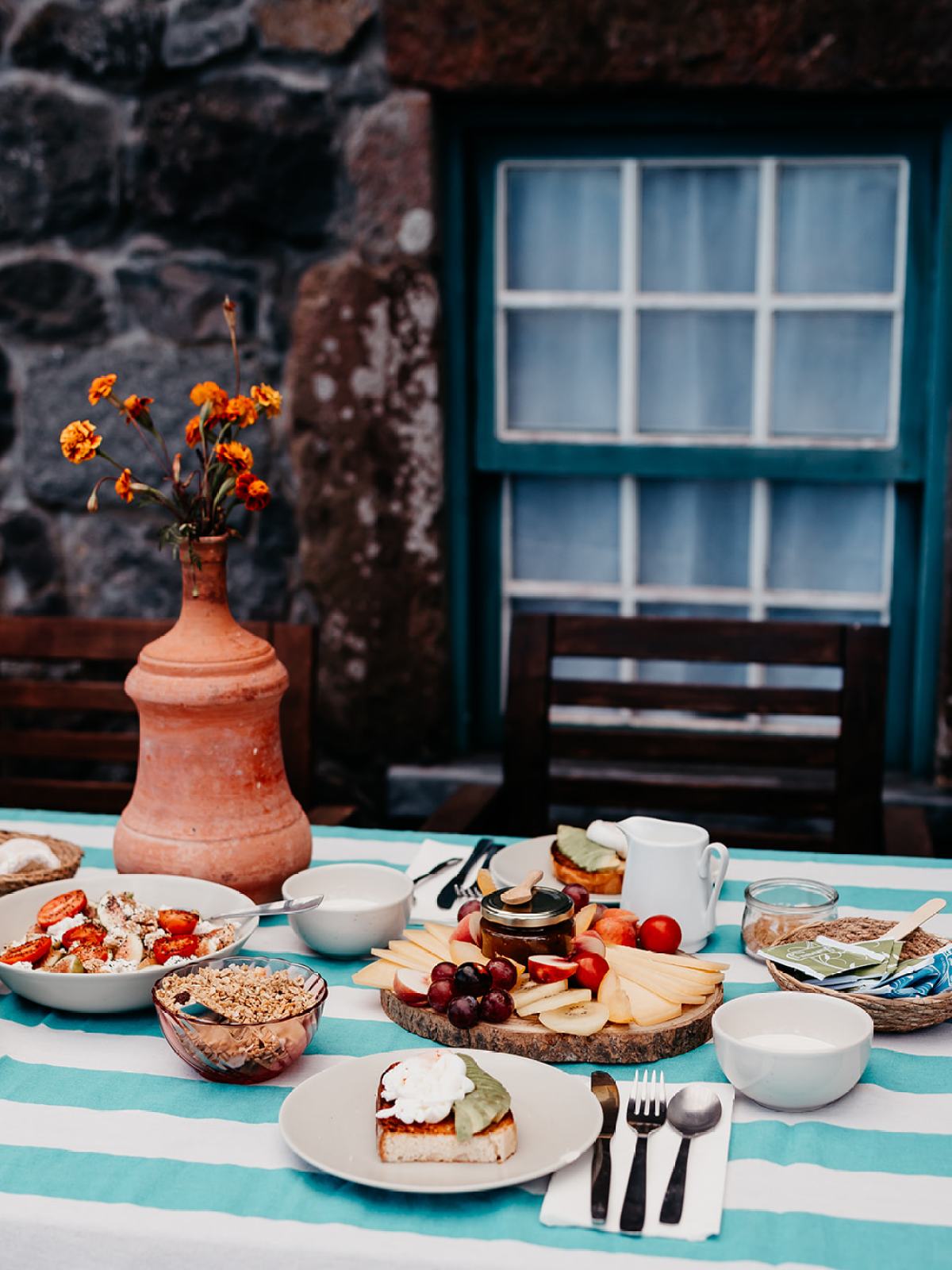 Outdoor table with brunch foods, striped cloth, flowers in vase, and stone wall backdrop.