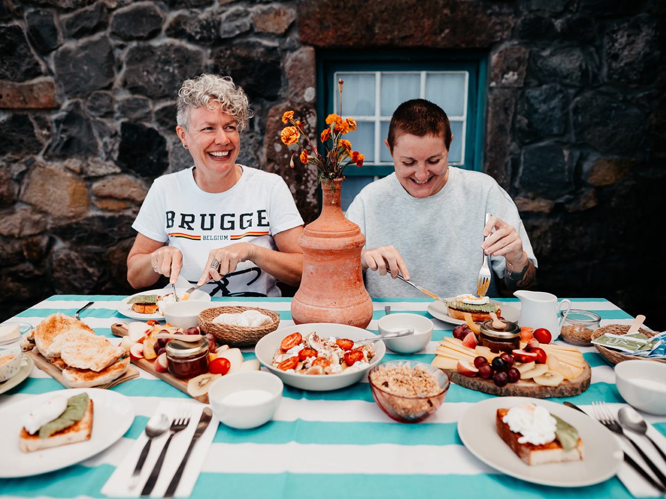 Two people enjoying an outdoor breakfast with various dishes on a blue-striped tablecloth.