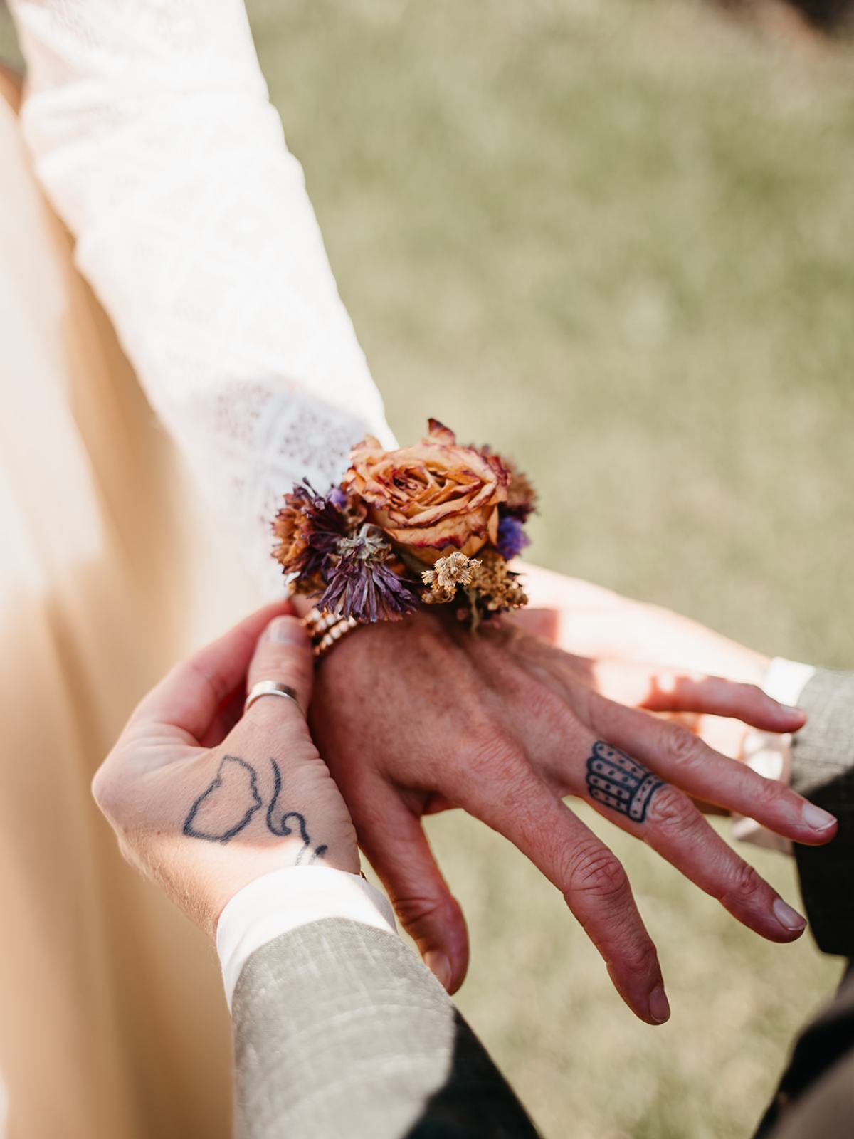 Tattooed hands adjusting a floral wrist corsage on a person's wrist outdoors.