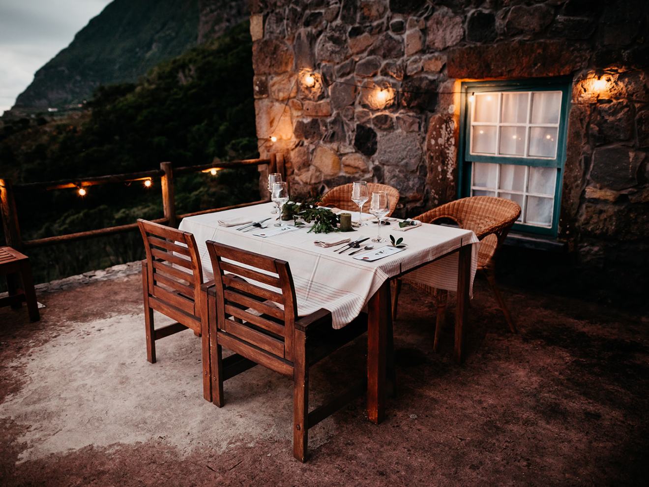 Rustic outdoor dining setup with wooden chairs, tablecloth, and string lights by a stone wall.
