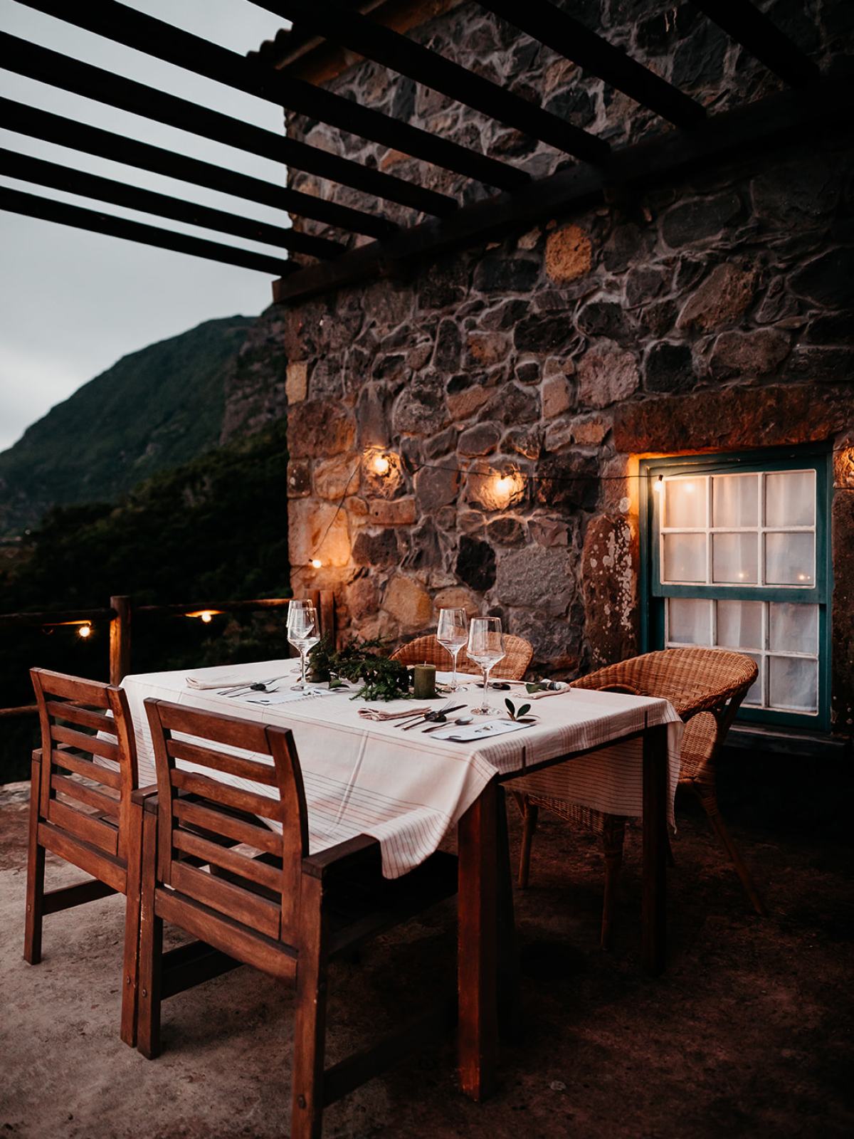 Outdoor dining table set for dinner against a stone wall with warm lighting.