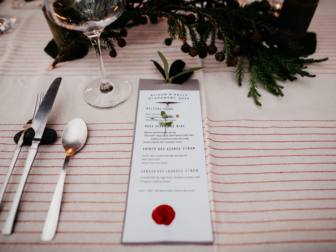 Dinner table setup with menu, cutlery, wine glass, and greenery on striped cloth.